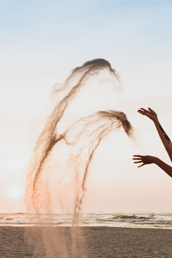 A person playfully tosses sand into the air at the beach during sunset, capturing freedom and motion.